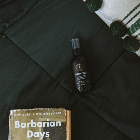 Bottle of Coastal Wolf Room Spray product on a dark surface with a book titled 'Barbarian Days' below.