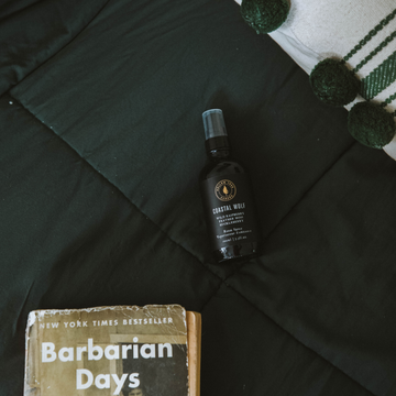 Bottle of Coastal Wolf Room Spray product on a dark surface with a book titled 'Barbarian Days' below.