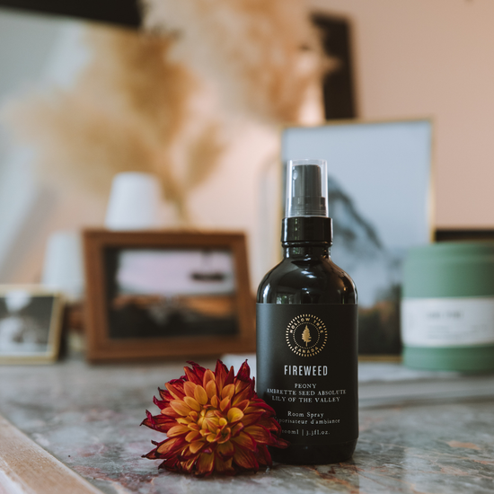 Room spray bottle with black label and silver mist sprayer, on tabletop with flower.