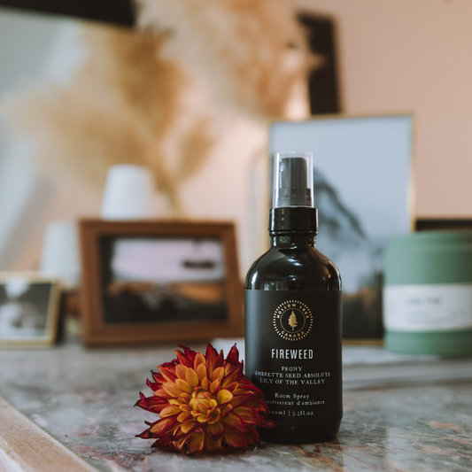 Room spray bottle with black label and silver mist sprayer, on tabletop with flower.