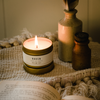 Lit travel candle in white tin with gold lid; wax visible in open top